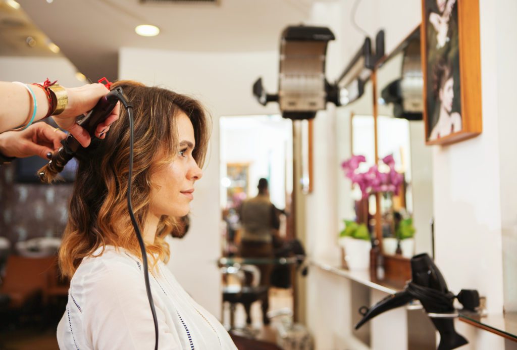 Hairdresser using curling tongs on customer's long brown hair in salon